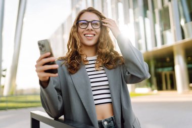 Portrait of stylish woman wearing glasses using phone against backdrop of modern office building. Beautiful businesswoman uses phone outdoors. Concepts of business, technology, and style.