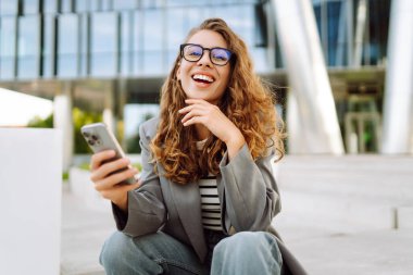 Portrait of stylish woman wearing glasses using phone against backdrop of modern office building. Beautiful businesswoman uses phone outdoors. Concepts of business, technology, and style.