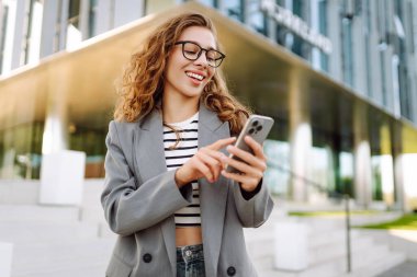 Portrait of stylish woman wearing glasses using phone against backdrop of modern office building. Beautiful businesswoman uses phone outdoors. Concepts of business, technology, and style.