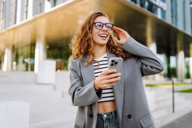 Portrait of stylish woman wearing glasses using phone against backdrop of modern office building. Beautiful businesswoman uses phone outdoors. Concepts of business, technology, and style.