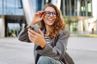 Portrait of stylish woman wearing glasses using phone against backdrop of modern office building. Beautiful businesswoman uses phone outdoors. Concepts of business, technology, and style.