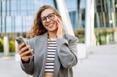 Portrait of stylish woman wearing glasses using phone against backdrop of modern office building. Beautiful businesswoman uses phone outdoors. Concepts of business, technology, and style.