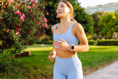 A beautiful woman in bright sportswear enjoys a jog in a sunny park. An athletic woman wearing wireless headphones runs outdoors in a tropical setting. Concept of sports, active lifestyle.