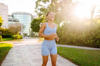 A beautiful woman in bright sportswear enjoys a jog in a sunny park. An athletic woman wearing wireless headphones runs outdoors in a tropical setting. Concept of sports, active lifestyle.