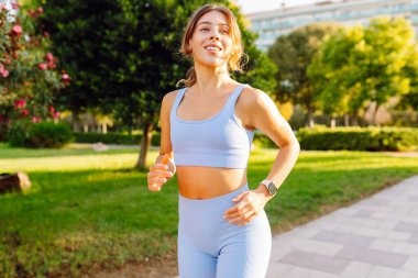 A beautiful woman in bright sportswear enjoys a jog in a sunny park. An athletic woman wearing wireless headphones runs outdoors in a tropical setting. Concept of sports, active lifestyle.