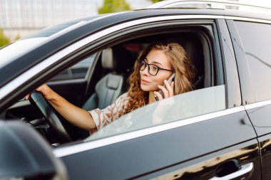 A young woman wearing glasses drives a black car on a sunny day. A beautiful woman behind the wheel of a car, holding a phone. Concept of transportation, urban life.