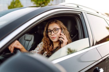 A young woman wearing glasses drives a black car on a sunny day. A beautiful woman behind the wheel of a car, holding a phone. Concept of transportation, urban life.