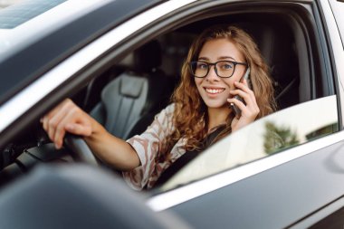 A young woman wearing glasses drives a black car on a sunny day. A beautiful woman behind the wheel of a car, holding a phone. Concept of transportation, urban life.