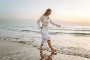 Beyaz etek ve üst giyen neşeli bir kadın gün batımında kumlu bir sahilde eğleniyor. Genç kadın deniz manzarasının tadını çıkarıyor ve kendini özgür hissediyor. Doğa kavramı, keyif.