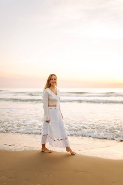 Beyaz etek ve üst giyen neşeli bir kadın gün batımında kumlu bir sahilde eğleniyor. Genç kadın deniz manzarasının tadını çıkarıyor ve kendini özgür hissediyor. Doğa kavramı, keyif.