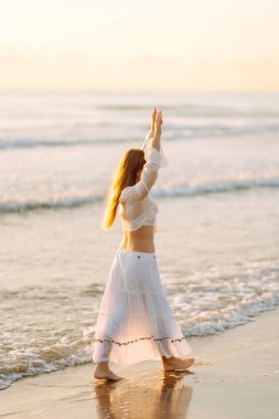 Beyaz etek ve üst giyen neşeli bir kadın gün batımında kumlu bir sahilde eğleniyor. Genç kadın deniz manzarasının tadını çıkarıyor ve kendini özgür hissediyor. Doğa kavramı, keyif.