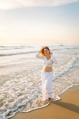 Beyaz etek ve üst giyen neşeli bir kadın gün batımında kumlu bir sahilde eğleniyor. Genç kadın deniz manzarasının tadını çıkarıyor ve kendini özgür hissediyor. Doğa kavramı, keyif.
