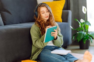 Kablosuz kulaklık takan genç bir kadın rahat oturma odasında otururken kitap okuyor. Güzel bir öğrenci kitap okur ve ev ortamında kanepenin yanına not yazar. Eğitim ve konfor kavramları.