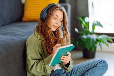 Kablosuz kulaklık takan genç bir kadın rahat oturma odasında otururken kitap okuyor. Güzel bir öğrenci kitap okur ve ev ortamında kanepenin yanına not yazar. Eğitim ve konfor kavramları.