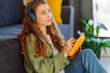 Kablosuz kulaklık takan genç bir kadın rahat oturma odasında otururken kitap okuyor. Güzel bir öğrenci kitap okur ve ev ortamında kanepenin yanına not yazar. Eğitim ve konfor kavramları.
