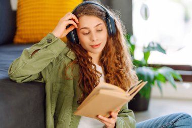 Kablosuz kulaklık takan genç bir kadın rahat oturma odasında otururken kitap okuyor. Güzel bir öğrenci kitap okur ve ev ortamında kanepenin yanına not yazar. Eğitim ve konfor kavramları.