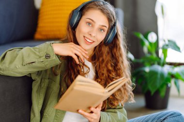 Kablosuz kulaklık takan genç bir kadın rahat oturma odasında otururken kitap okuyor. Güzel bir öğrenci kitap okur ve ev ortamında kanepenin yanına not yazar. Eğitim ve konfor kavramları.