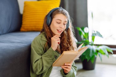Kablosuz kulaklık takan genç bir kadın rahat oturma odasında otururken kitap okuyor. Güzel bir öğrenci kitap okur ve ev ortamında kanepenin yanına not yazar. Eğitim ve konfor kavramları.