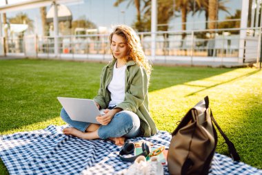 Dizüstü bilgisayarı olan gülümseyen bir kadın güneşli parkta, palmiye ağaçlarıyla çevrili bir şekilde oturuyor. Serbest çalışan kadın dizüstü bilgisayarda çalışıyor ve gün batımını dışarıda izlemeyi seviyor. Konsept olarak uzak çalışma, hafta sonları ya da eğitim için.