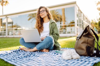 Dizüstü bilgisayarı olan gülümseyen bir kadın güneşli parkta, palmiye ağaçlarıyla çevrili bir şekilde oturuyor. Serbest çalışan kadın dizüstü bilgisayarda çalışıyor ve gün batımını dışarıda izlemeyi seviyor. Konsept olarak uzak çalışma, hafta sonları ya da eğitim için.