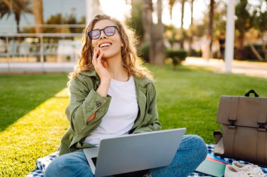 Dizüstü bilgisayarı olan gülümseyen bir kadın güneşli parkta, palmiye ağaçlarıyla çevrili bir şekilde oturuyor. Serbest çalışan kadın dizüstü bilgisayarda çalışıyor ve gün batımını dışarıda izlemeyi seviyor. Konsept olarak uzak çalışma, hafta sonları ya da eğitim için.