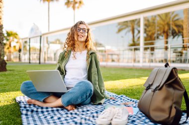 Dizüstü bilgisayarı olan gülümseyen bir kadın güneşli parkta, palmiye ağaçlarıyla çevrili bir şekilde oturuyor. Serbest çalışan kadın dizüstü bilgisayarda çalışıyor ve gün batımını dışarıda izlemeyi seviyor. Konsept olarak uzak çalışma, hafta sonları ya da eğitim için.