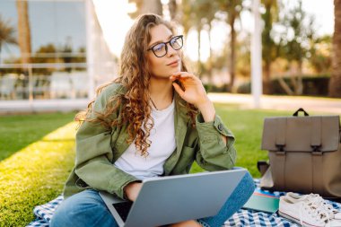 Dizüstü bilgisayarı olan gülümseyen bir kadın güneşli parkta, palmiye ağaçlarıyla çevrili bir şekilde oturuyor. Serbest çalışan kadın dizüstü bilgisayarda çalışıyor ve gün batımını dışarıda izlemeyi seviyor. Konsept olarak uzak çalışma, hafta sonları ya da eğitim için.