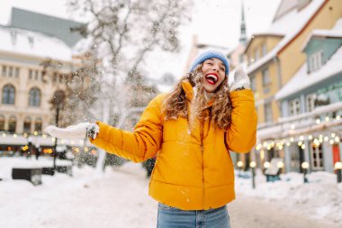 Şehrin sokaklarında karda eğlenen mutlu bir kadının portresi. Parlak ceketli güzel bir turist şehrin sokaklarında kış gününün tadını çıkarıyor. Turizm ve eğlence kavramı.