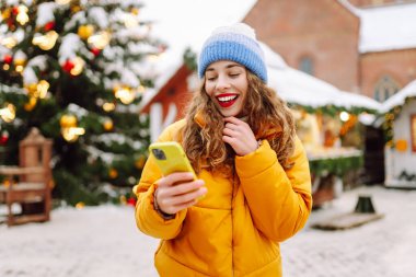 Parlak ceketli ve telefonlu mutlu bir kadın. Parlak ışıklarla Noel pazarının arka planında duruyor. Güzel turistler telefon ve neşeli Noel atmosferiyle güneşli bir kış gününün tadını çıkarıyorlar.. 
