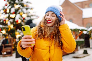 Parlak ceketli ve telefonlu mutlu bir kadın. Parlak ışıklarla Noel pazarının arka planında duruyor. Güzel turistler telefon ve neşeli Noel atmosferiyle güneşli bir kış gününün tadını çıkarıyorlar.. 