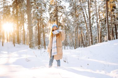 Sıcak kış kıyafetleri içinde neşeli bir kadın güneşli bir ormanda karla eğlenir. Güzel bir kadın dışarıda kış gününün tadını çıkarır. Eğlence anlayışı, doğa. Etkin yaşam biçimi.