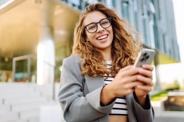 Portrait of stylish woman wearing glasses using phone against backdrop of modern office building. Beautiful businesswoman uses phone outdoors. Concepts of business, technology, and style.