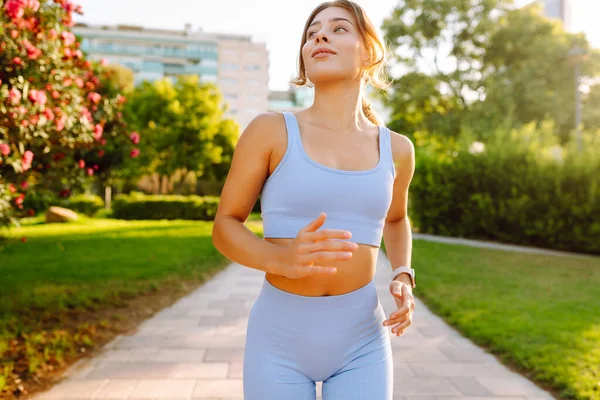 A beautiful woman in bright sportswear enjoys a jog in a sunny park. An athletic woman wearing wireless headphones runs outdoors in a tropical setting. Concept of sports, active lifestyle.