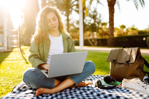 Dizüstü bilgisayarı olan gülümseyen bir kadın güneşli parkta, palmiye ağaçlarıyla çevrili bir şekilde oturuyor. Serbest çalışan kadın dizüstü bilgisayarda çalışıyor ve gün batımını dışarıda izlemeyi seviyor. Konsept olarak uzak çalışma, hafta sonları ya da eğitim için.