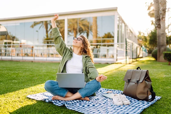 Dizüstü bilgisayarı olan gülümseyen bir kadın güneşli parkta, palmiye ağaçlarıyla çevrili bir şekilde oturuyor. Serbest çalışan kadın dizüstü bilgisayarda çalışıyor ve gün batımını dışarıda izlemeyi seviyor. Konsept olarak uzak çalışma, hafta sonları ya da eğitim için.