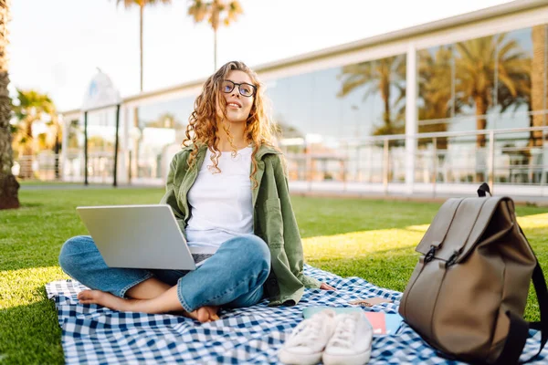 Dizüstü bilgisayarı olan gülümseyen bir kadın güneşli parkta, palmiye ağaçlarıyla çevrili bir şekilde oturuyor. Serbest çalışan kadın dizüstü bilgisayarda çalışıyor ve gün batımını dışarıda izlemeyi seviyor. Konsept olarak uzak çalışma, hafta sonları ya da eğitim için.