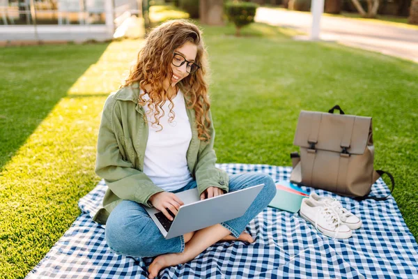 Dizüstü bilgisayarı olan gülümseyen bir kadın güneşli parkta, palmiye ağaçlarıyla çevrili bir şekilde oturuyor. Serbest çalışan kadın dizüstü bilgisayarda çalışıyor ve gün batımını dışarıda izlemeyi seviyor. Konsept olarak uzak çalışma, hafta sonları ya da eğitim için.