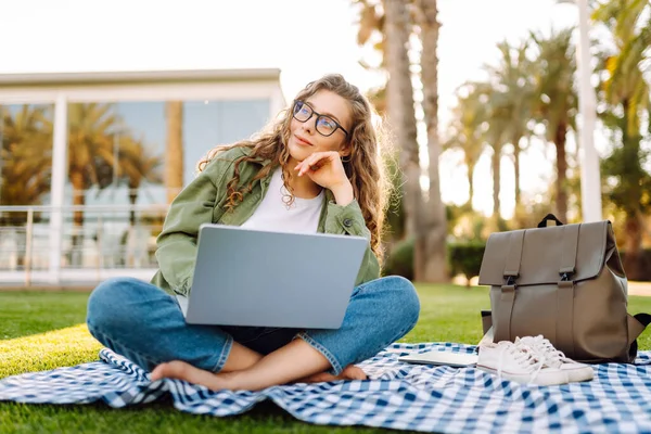 Dizüstü bilgisayarı olan gülümseyen bir kadın güneşli parkta, palmiye ağaçlarıyla çevrili bir şekilde oturuyor. Serbest çalışan kadın dizüstü bilgisayarda çalışıyor ve gün batımını dışarıda izlemeyi seviyor. Konsept olarak uzak çalışma, hafta sonları ya da eğitim için.