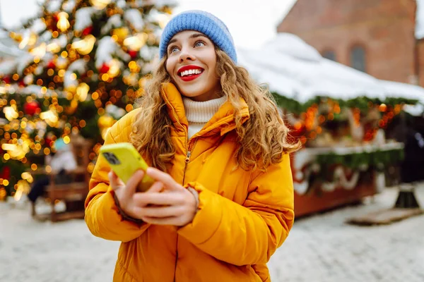 Parlak ceketli ve telefonlu mutlu bir kadın. Parlak ışıklarla Noel pazarının arka planında duruyor. Güzel turistler telefon ve neşeli Noel atmosferiyle güneşli bir kış gününün tadını çıkarıyorlar.. 