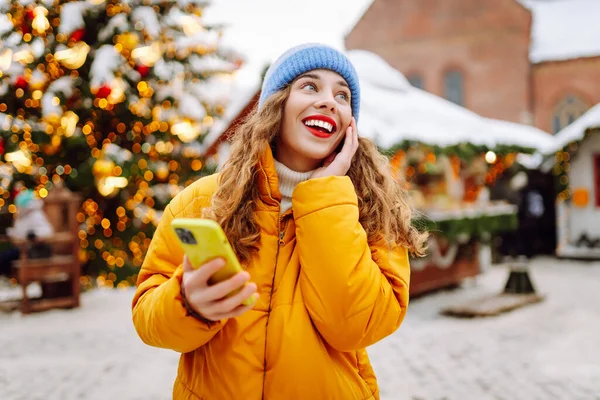 Parlak ceketli ve telefonlu mutlu bir kadın. Parlak ışıklarla Noel pazarının arka planında duruyor. Güzel turistler telefon ve neşeli Noel atmosferiyle güneşli bir kış gününün tadını çıkarıyorlar.. 