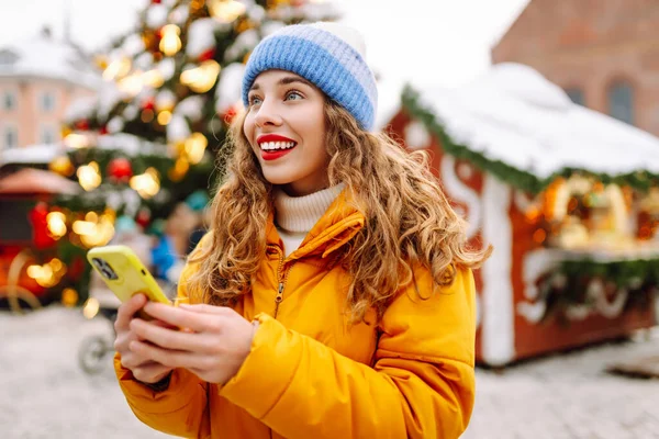 Parlak ceketli ve telefonlu mutlu bir kadın. Parlak ışıklarla Noel pazarının arka planında duruyor. Güzel turistler telefon ve neşeli Noel atmosferiyle güneşli bir kış gününün tadını çıkarıyorlar.. 