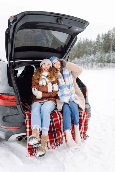 Sıcak kıyafetler içindeki iki neşeli kadın kış günü bir arabanın bagajında battaniyenin üzerinde otururlar. Güzel arkadaşlar, karlı bir ormanda araba yolculuğunun tadını çıkarırlar. Arkadaşlık, eğlence ve mevsimlik kavramları.