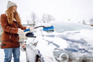Gülümseyen bir kadın karlı bir günde kar fırçasıyla arabasındaki karı temizler. Güzel bir kadın sürücü güneşli bir günde arabasındaki karı temizler. Buz kavramı, ulaşım. Soğuk bir gün..