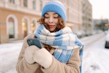 Parlak eşarplı ve şapkalı neşeli bir kadın telefonuyla karlı bir caddede duruyor. Güzel bir kadın telefonunda blog yazıyor, kış günü yürüyüş yapıyor. Günlük konsepti, kentsel.