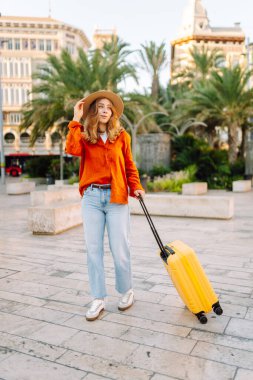 Sarı valizli, sırt çantalı ve güneşli meydanda yürüyen kadın bir gezginin dikiz görüntüsü. Güzel bir kadın küçük bir bavulla eski kasabanın sokaklarında dolaşıyor. Konsept: seyahat, turizm.