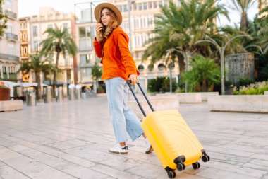 Sarı valizli, sırt çantalı ve güneşli meydanda yürüyen kadın bir gezginin dikiz görüntüsü. Güzel bir kadın küçük bir bavulla eski kasabanın sokaklarında dolaşıyor. Konsept: seyahat, turizm.