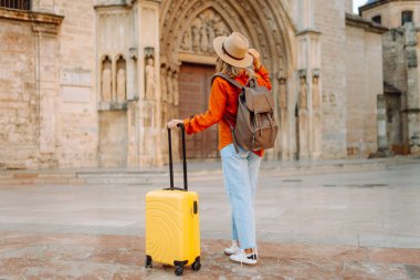 Sarı valizli, sırt çantalı ve güneşli meydanda yürüyen kadın bir gezginin dikiz görüntüsü. Güzel bir kadın küçük bir bavulla eski kasabanın sokaklarında dolaşıyor. Konsept: seyahat, turizm.