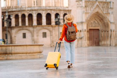 Sarı valizli, sırt çantalı ve güneşli meydanda yürüyen kadın bir gezginin dikiz görüntüsü. Güzel bir kadın küçük bir bavulla eski kasabanın sokaklarında dolaşıyor. Konsept: seyahat, turizm.