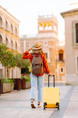 Sarı valizli, sırt çantalı ve güneşli meydanda yürüyen kadın bir gezginin dikiz görüntüsü. Güzel bir kadın küçük bir bavulla eski kasabanın sokaklarında dolaşıyor. Konsept: seyahat, turizm.