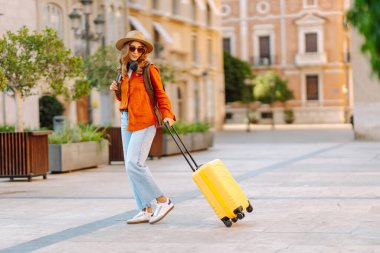 Sarı valizli, sırt çantalı ve güneşli meydanda yürüyen kadın bir gezginin dikiz görüntüsü. Güzel bir kadın küçük bir bavulla eski kasabanın sokaklarında dolaşıyor. Konsept: seyahat, turizm.